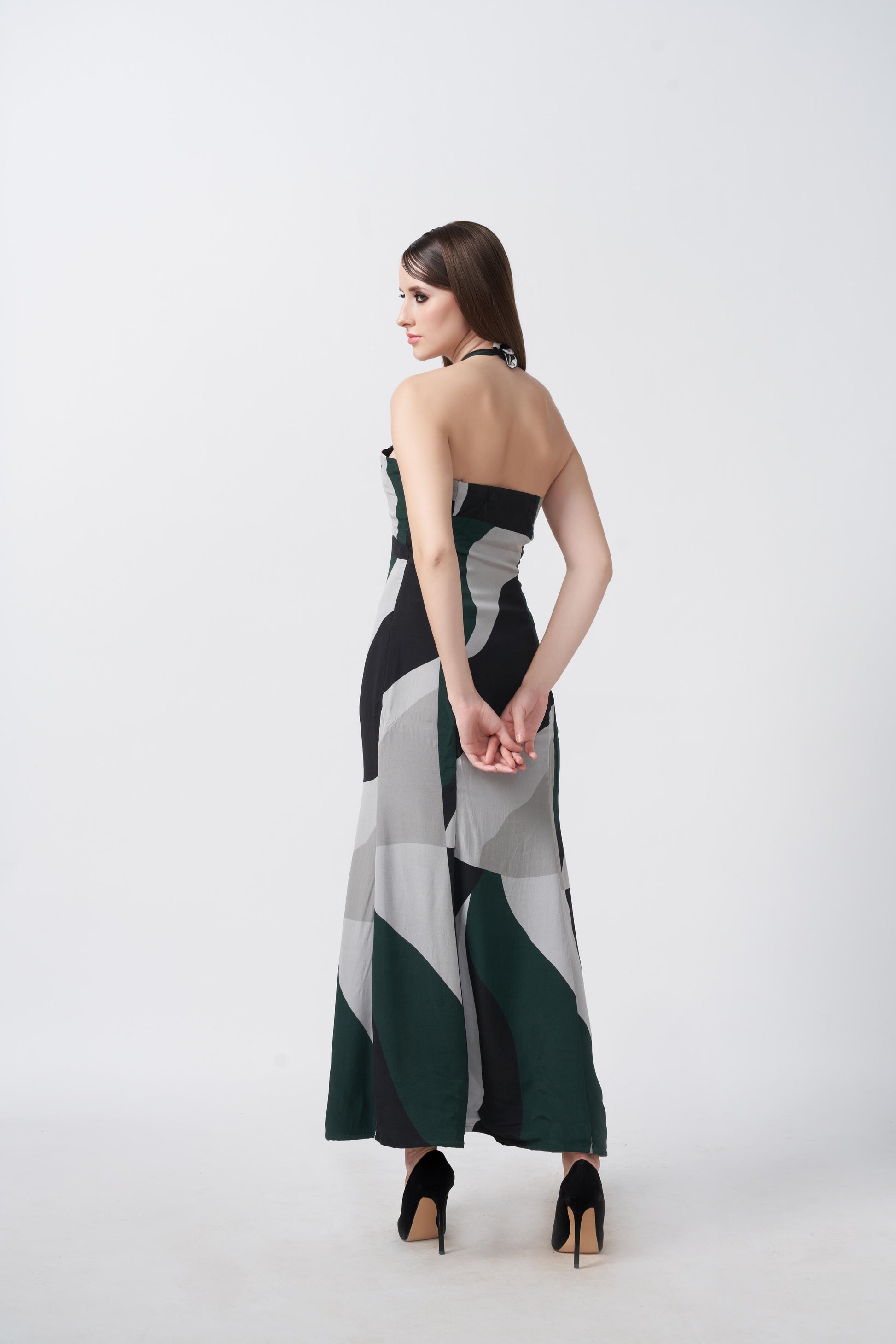 Rear view of model walking in green and grey abstract printed halter dress with tie-back and ankle length cut.