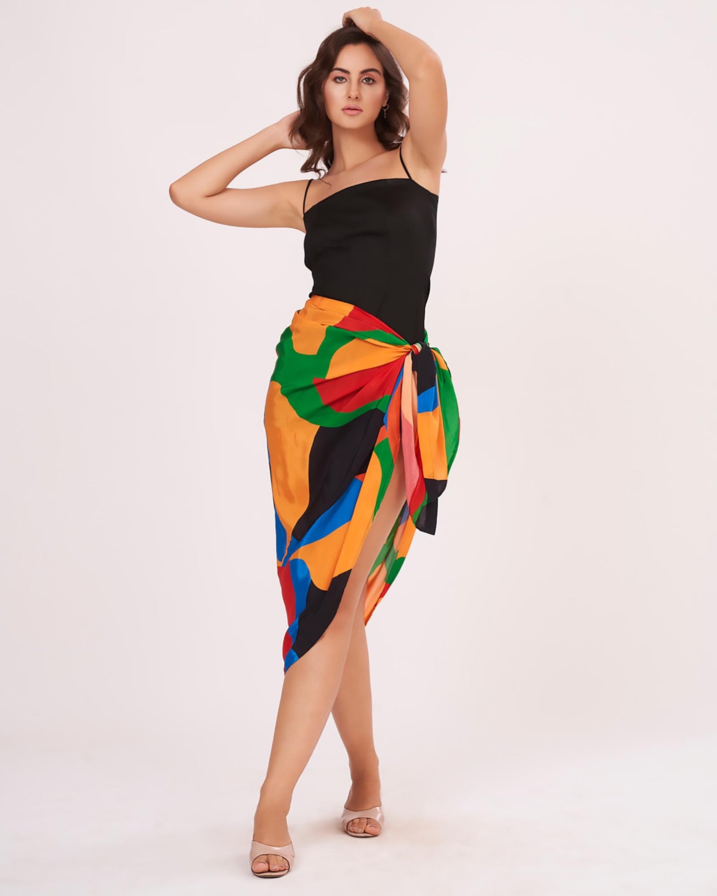 Model mid-walk with one arm raised, showing the side draping of the colorful sarong wrap paired with the fitted black dress.