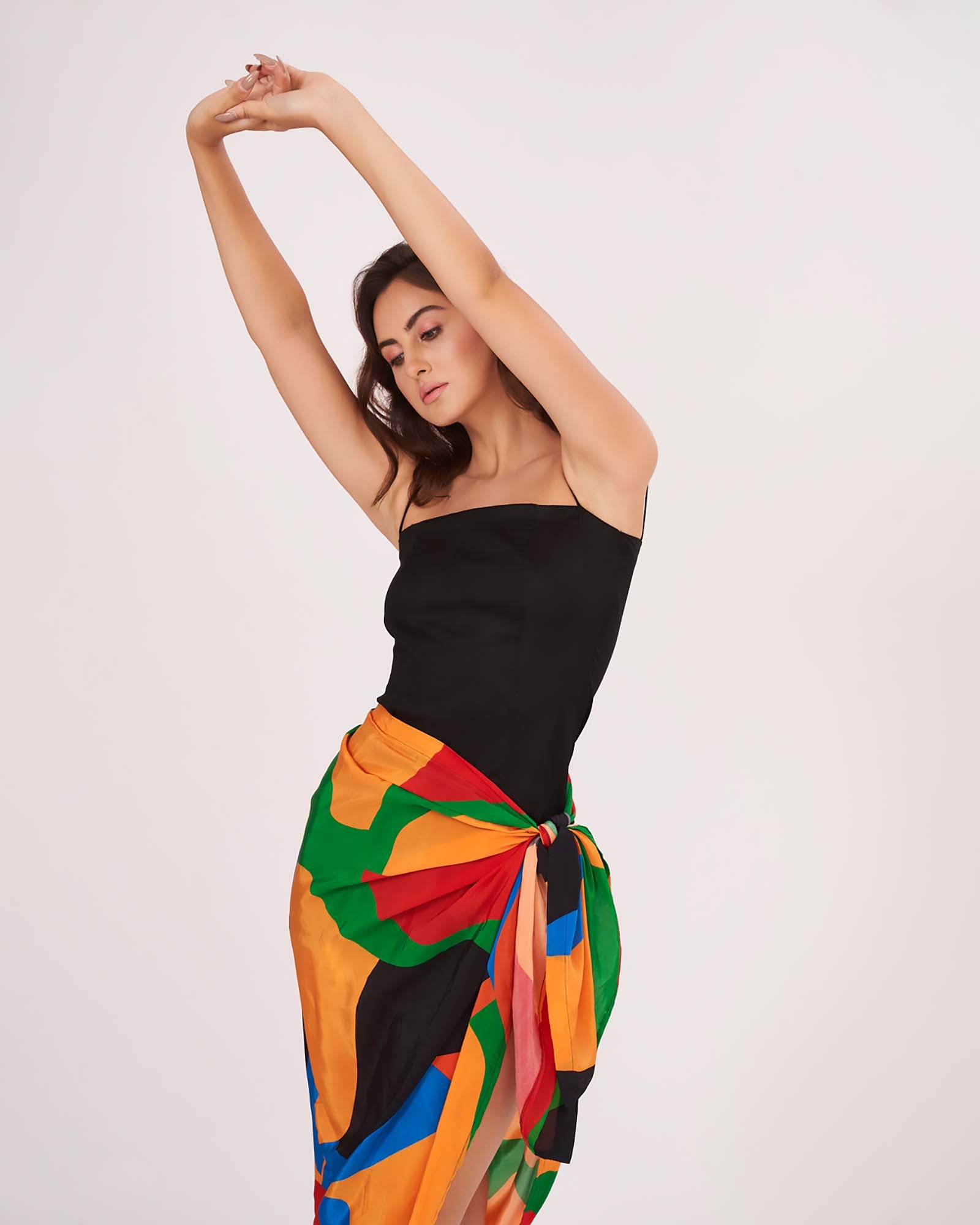 Playful standing pose with both arms overhead, showcasing the fitted silhouette of the black dress and asymmetric sarong tie-up.