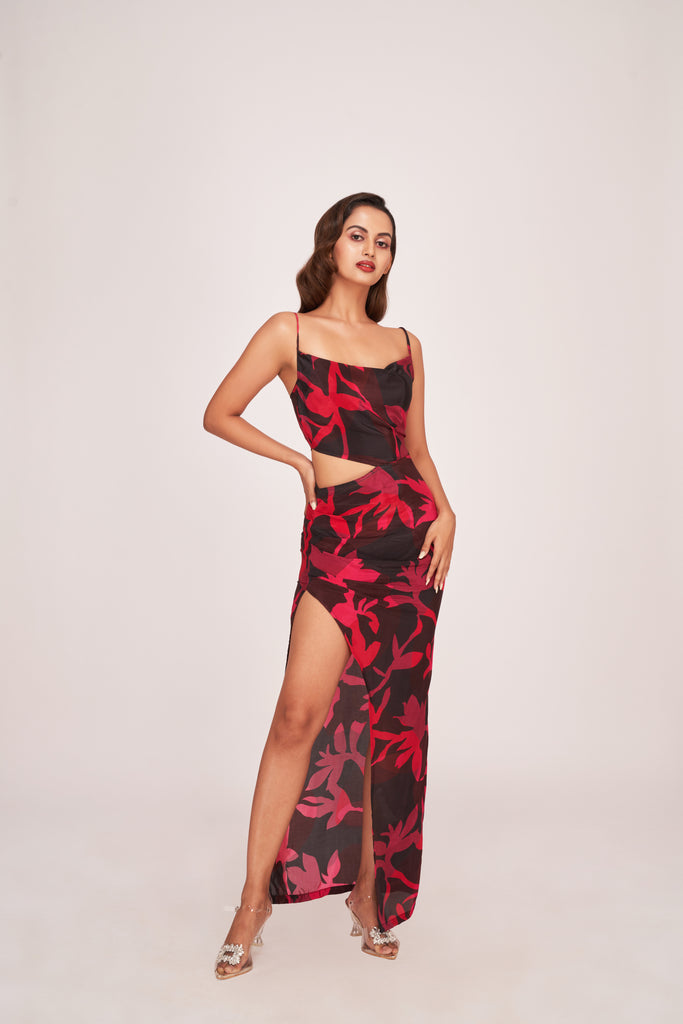 Model wearing a black floral ankle-length dress with cowl neck and thigh-high slit, posing with hand on hip.