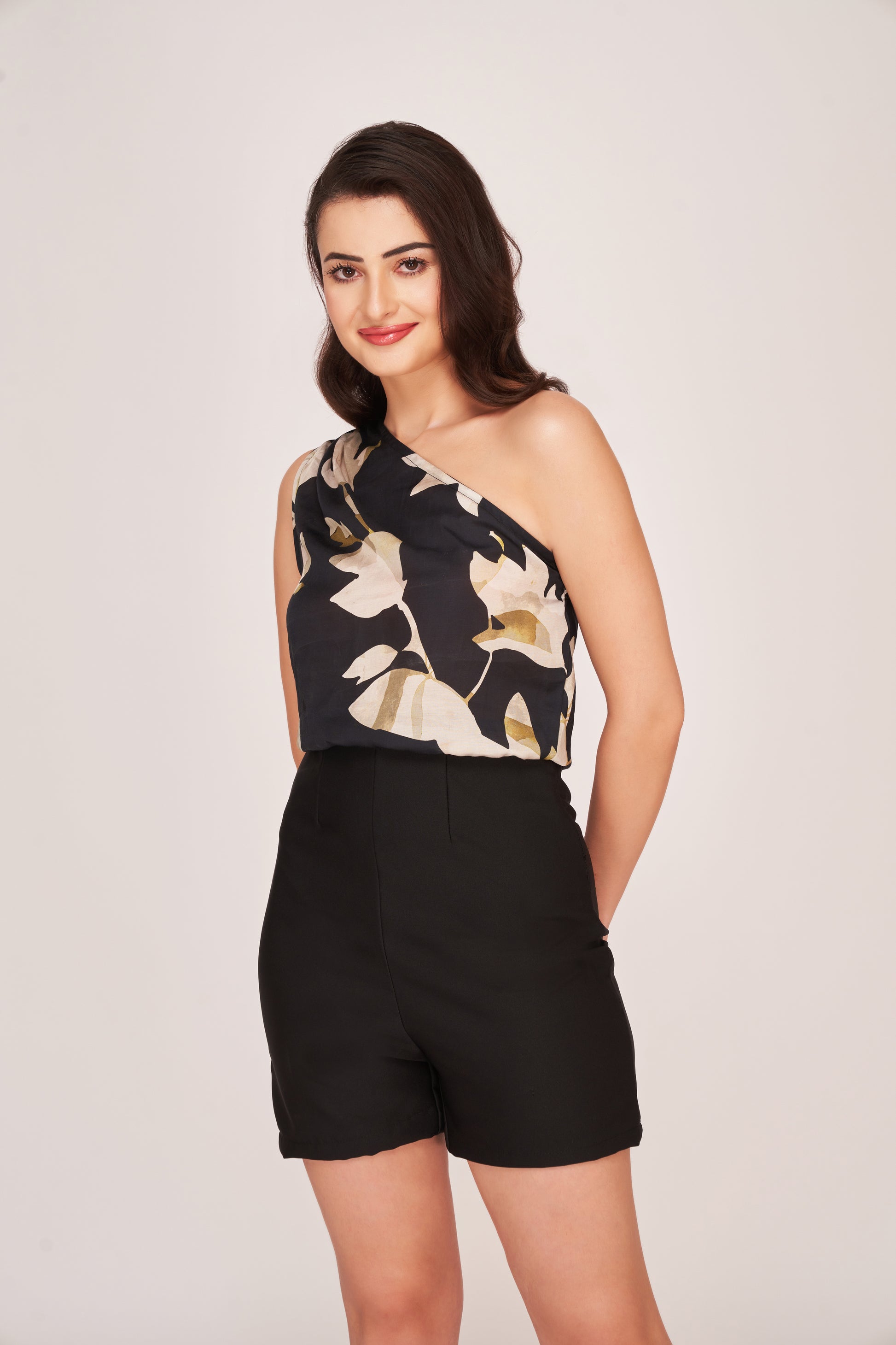 Model in a straight relaxed pose wearing a black floral one-shoulder top and high-waist shorts, highlighting the elegant asymmetry and clean silhouette.
