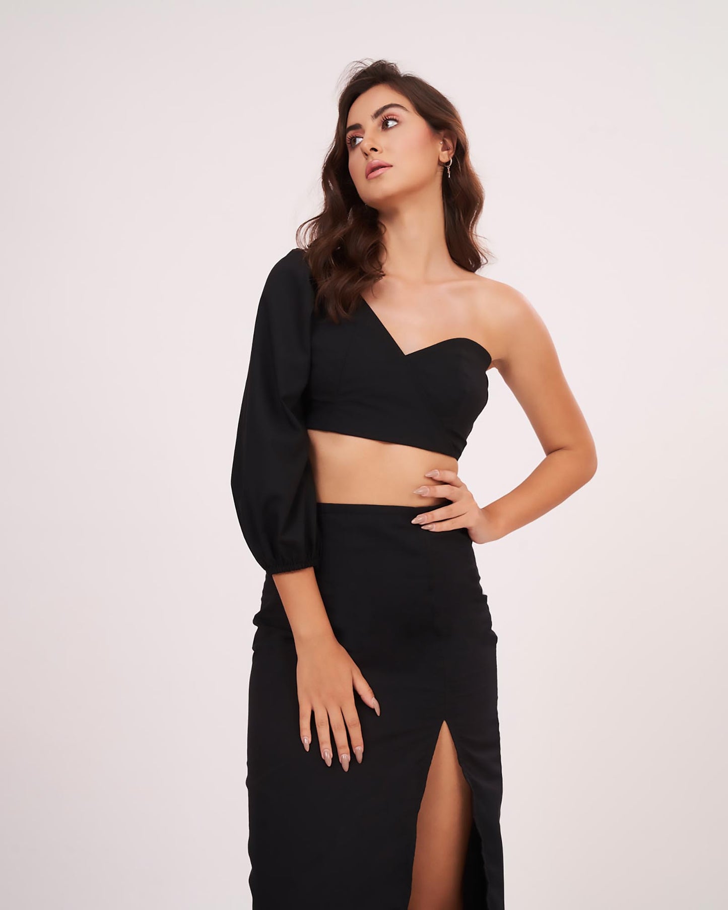 Model with hand on waist, showcasing the black one-sleeve crop top and thigh-high slit skirt.