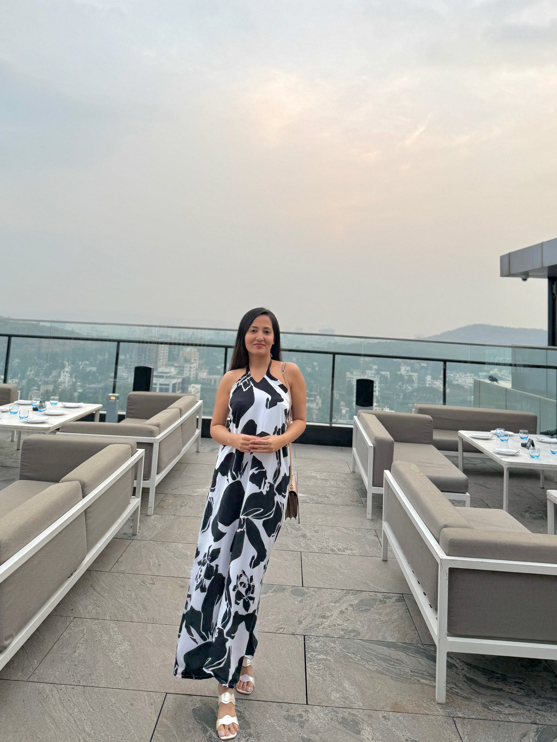Model standing on a rooftop in black and white abstract halter dress, facing camera with city view behind.