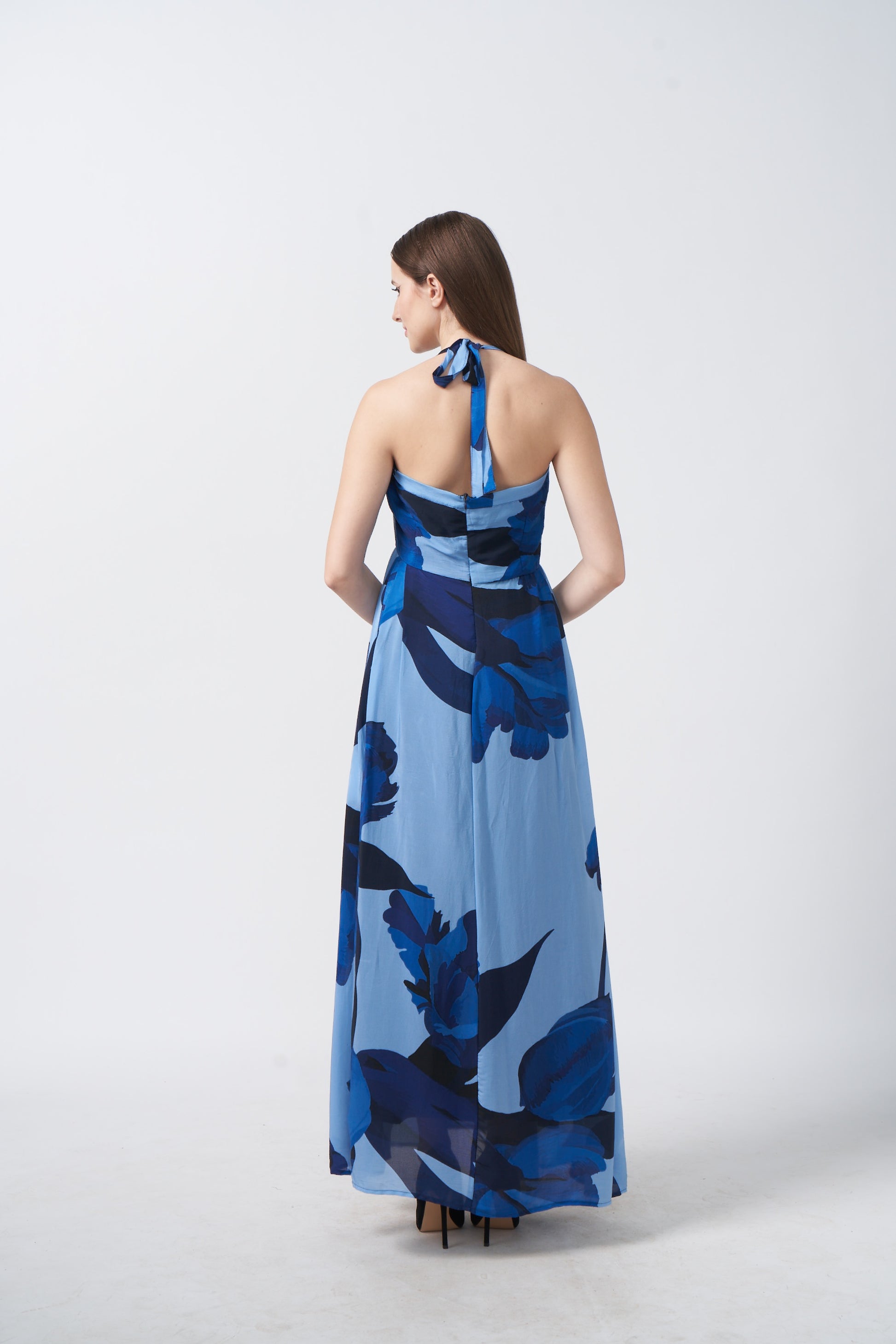 Back view of model in blue floral crepe halter dress, featuring tie-back neckline and fluid drape.