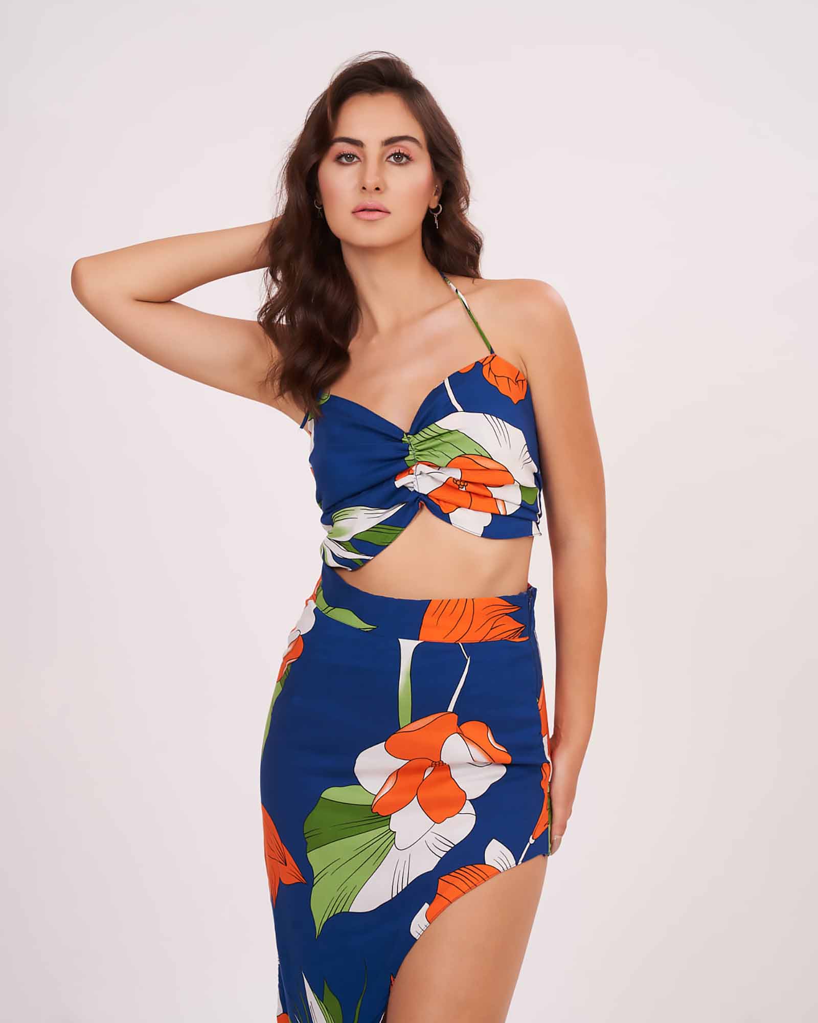 Side-turned pose with one hand raised to head, showing the ruched halter neckline and flowy slit of the floral skirt in the blue co-ord set.