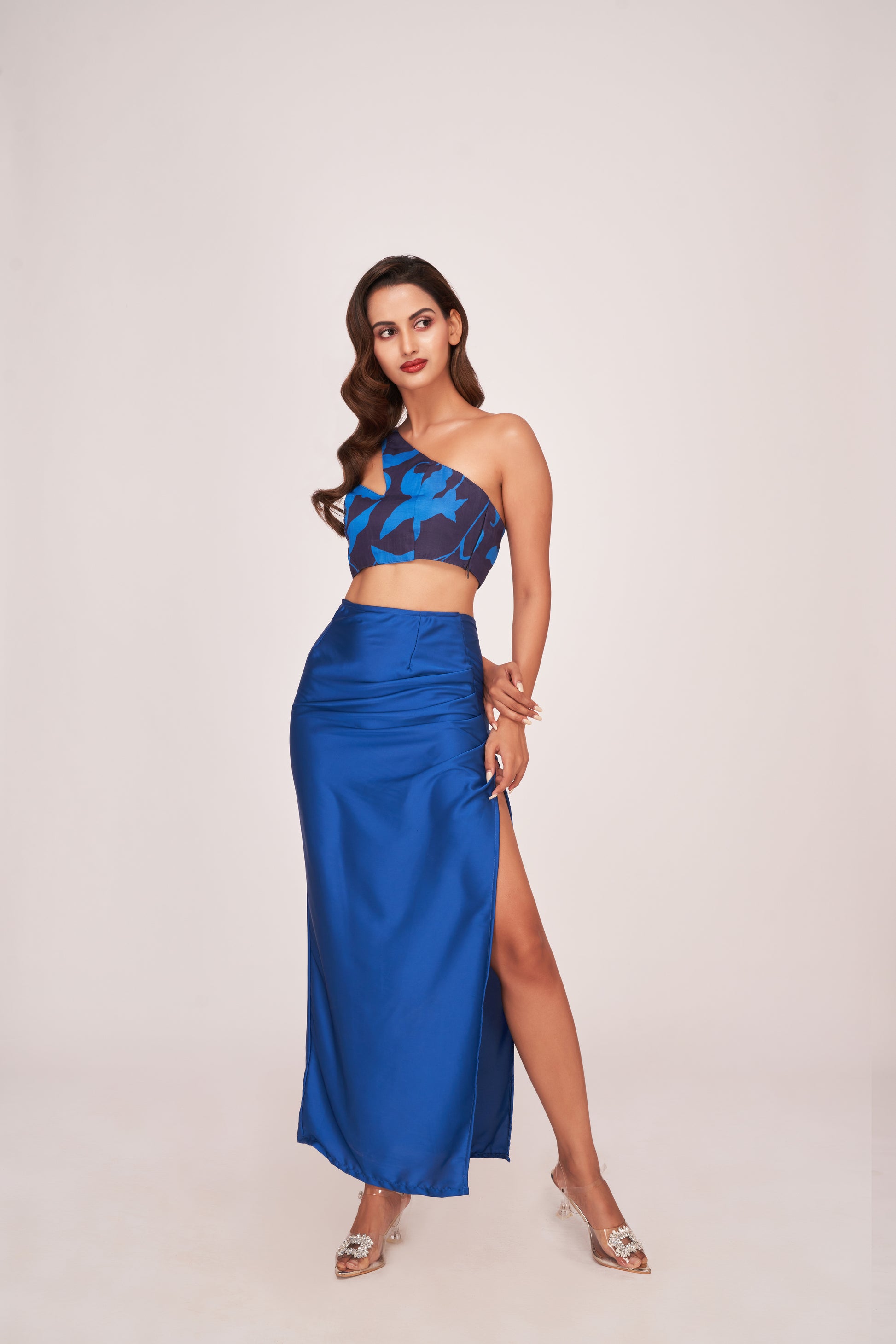Model standing straight in a satin blue co-ord set, showcasing the thigh-high slit and one-shoulder printed crop top.
