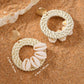 Shell Ring Drop Earrings in white featuring cowrie shells and woven straw hoops with measurement labels of 6.0cm by 4.8cm on a sand background.