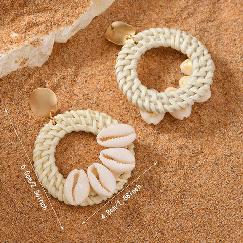 Shell Ring Drop Earrings in white featuring cowrie shells and woven straw hoops with measurement labels of 6.0cm by 4.8cm on a sand background.
