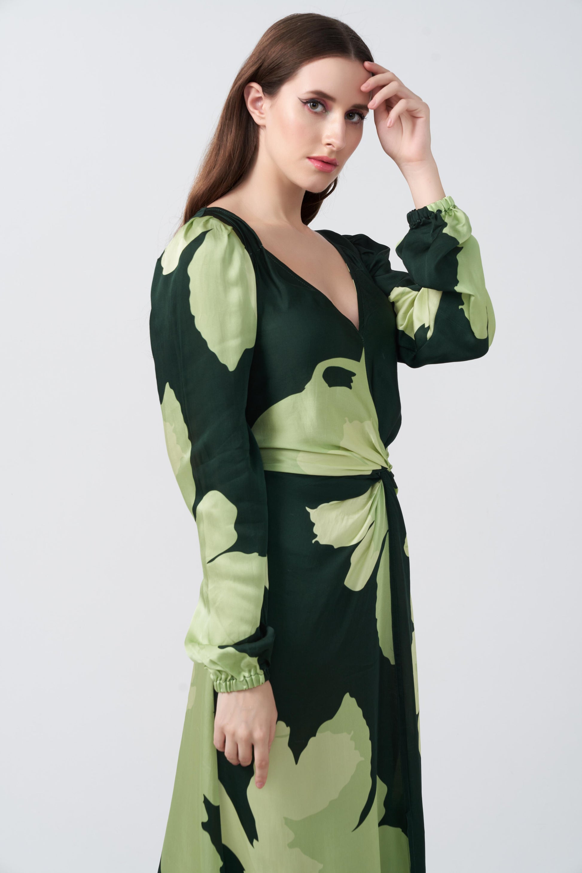 Model in profile pose with hand near face, showcasing the balloon sleeve and elegant V-neckline of the green floral satin dress.