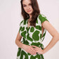 Close-up of model in green abstract sleeveless muslin dress, smiling with hands on waist and wrap neckline visible.