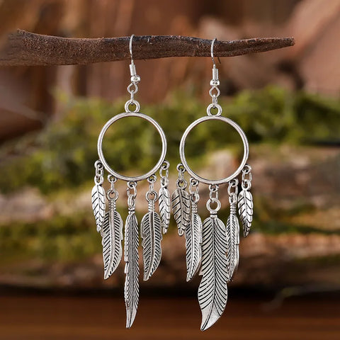 A pair of antique silver Feather Hoop Drop Earrings with intricate metallic feather charms hanging from a circular hoop, displayed on a rustic branch background.