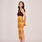 Model in a playful angled pose with both hands behind her back, wearing maroon and yellow abstract satin midi dress.