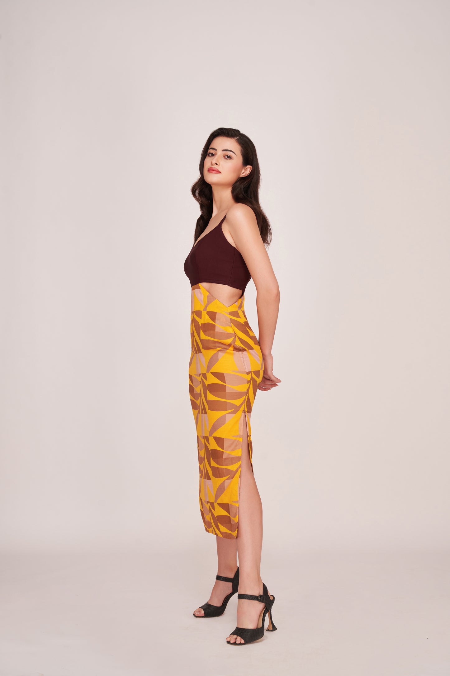 Model in a playful angled pose with both hands behind her back, wearing maroon and yellow abstract satin midi dress.