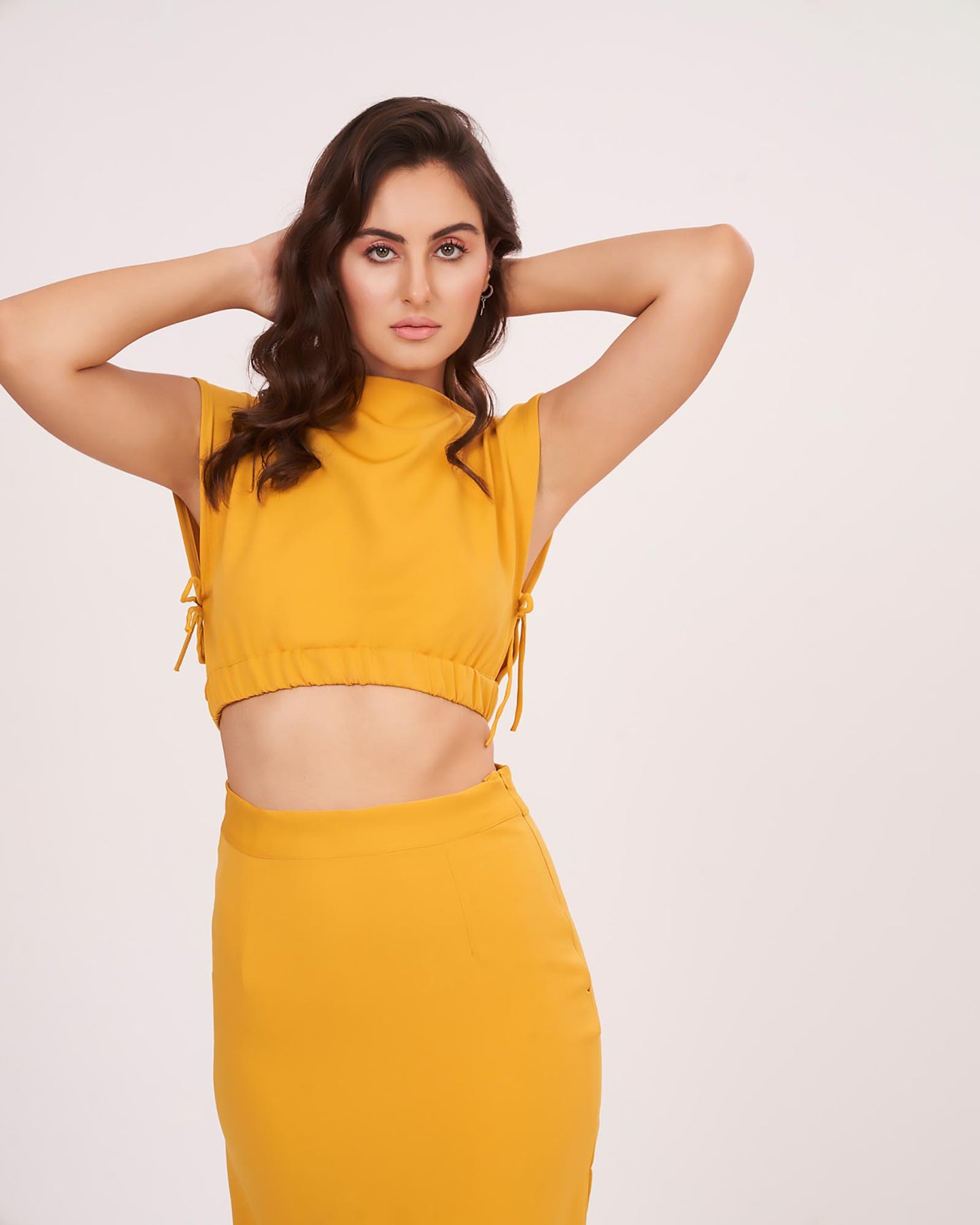 Model poses with hands behind her head, drawing attention to the clean lines of the sleeveless blouse and the confident styling of the look.