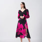 Model wearing a pink and black floral silk midi dress with V-neckline, long sleeves, and side slit, standing front-facing.
