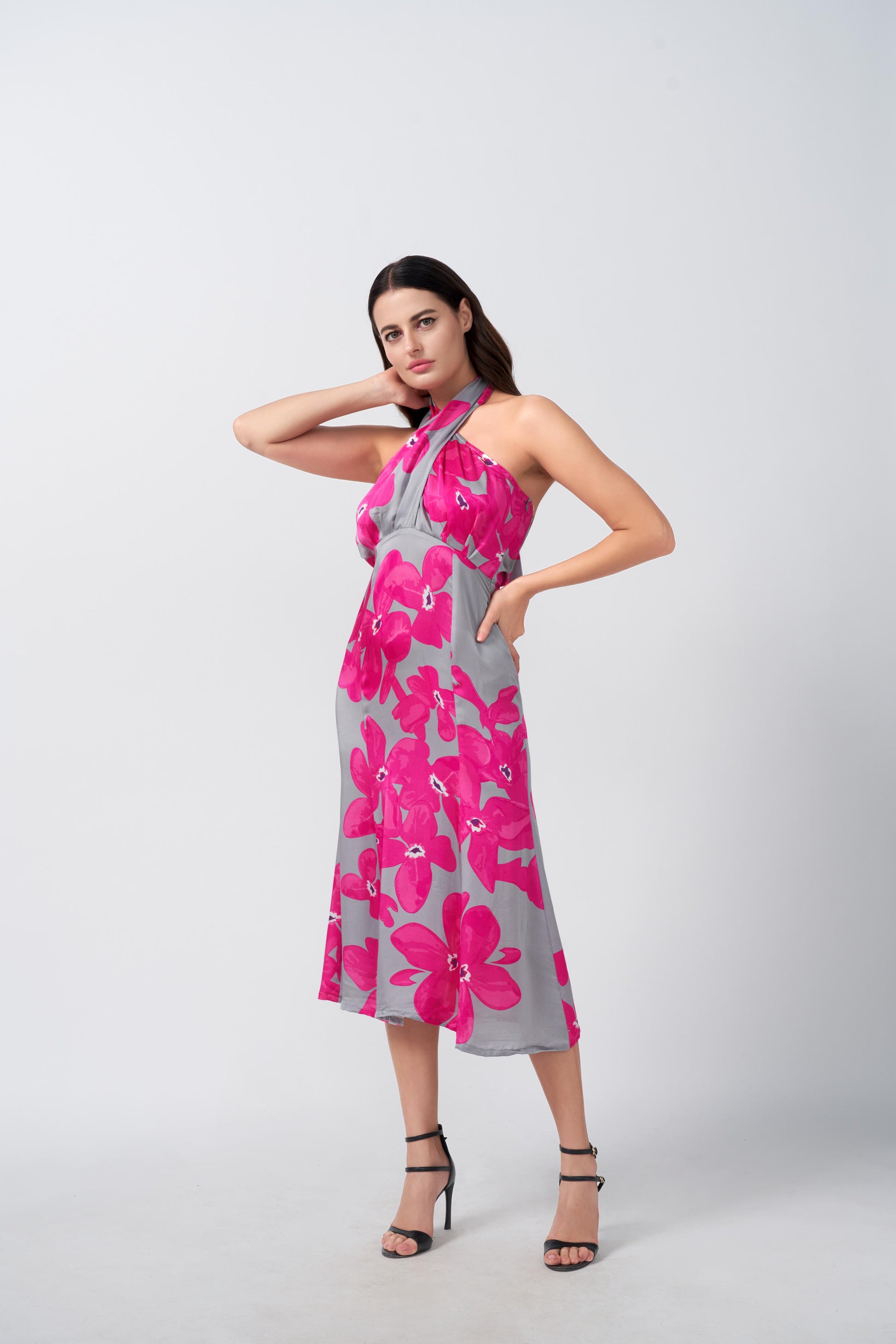 Model in motion with hand on hip, showcasing side flow of pink and grey floral midi dress and tie-back halter design.