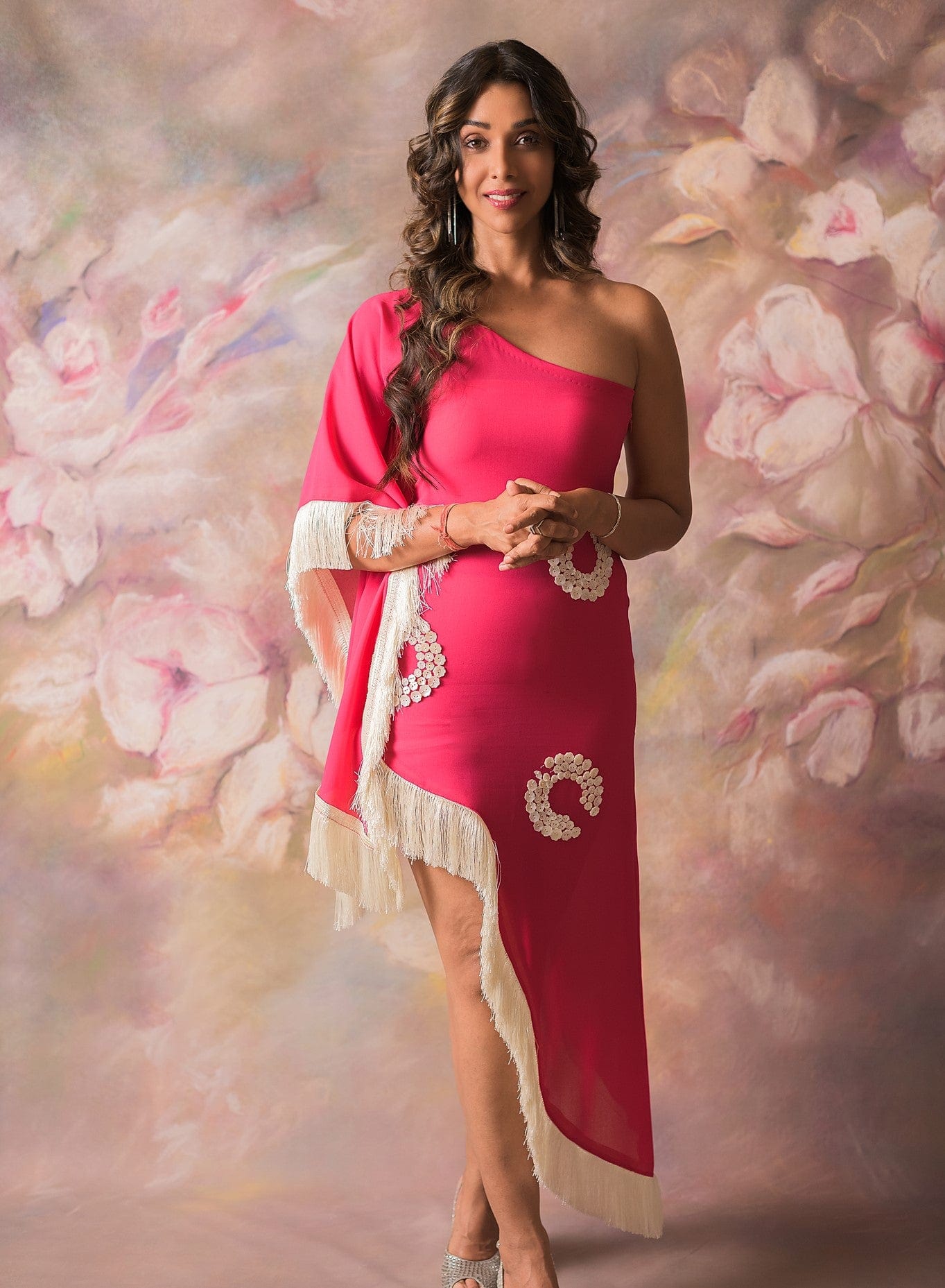 Model poses in front view wearing a pink one-shoulder georgette dress featuring shell-detailed embroidery and fringe hem, styled for a beach vacation or party night.