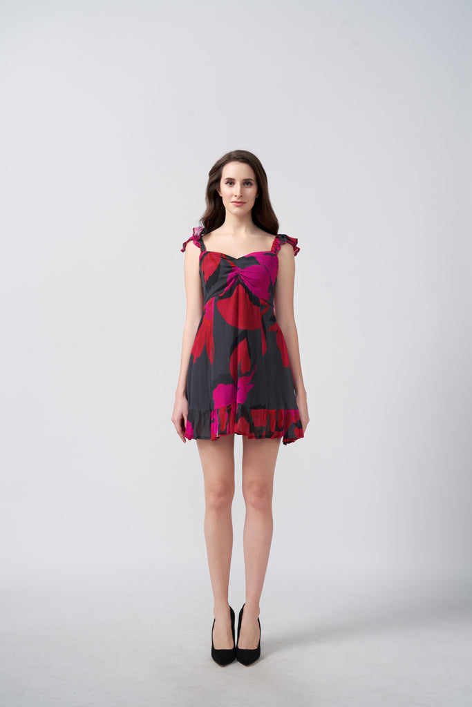 Model wearing a short red and pink abstract floral dress with frill shoulder straps, standing against a plain background.