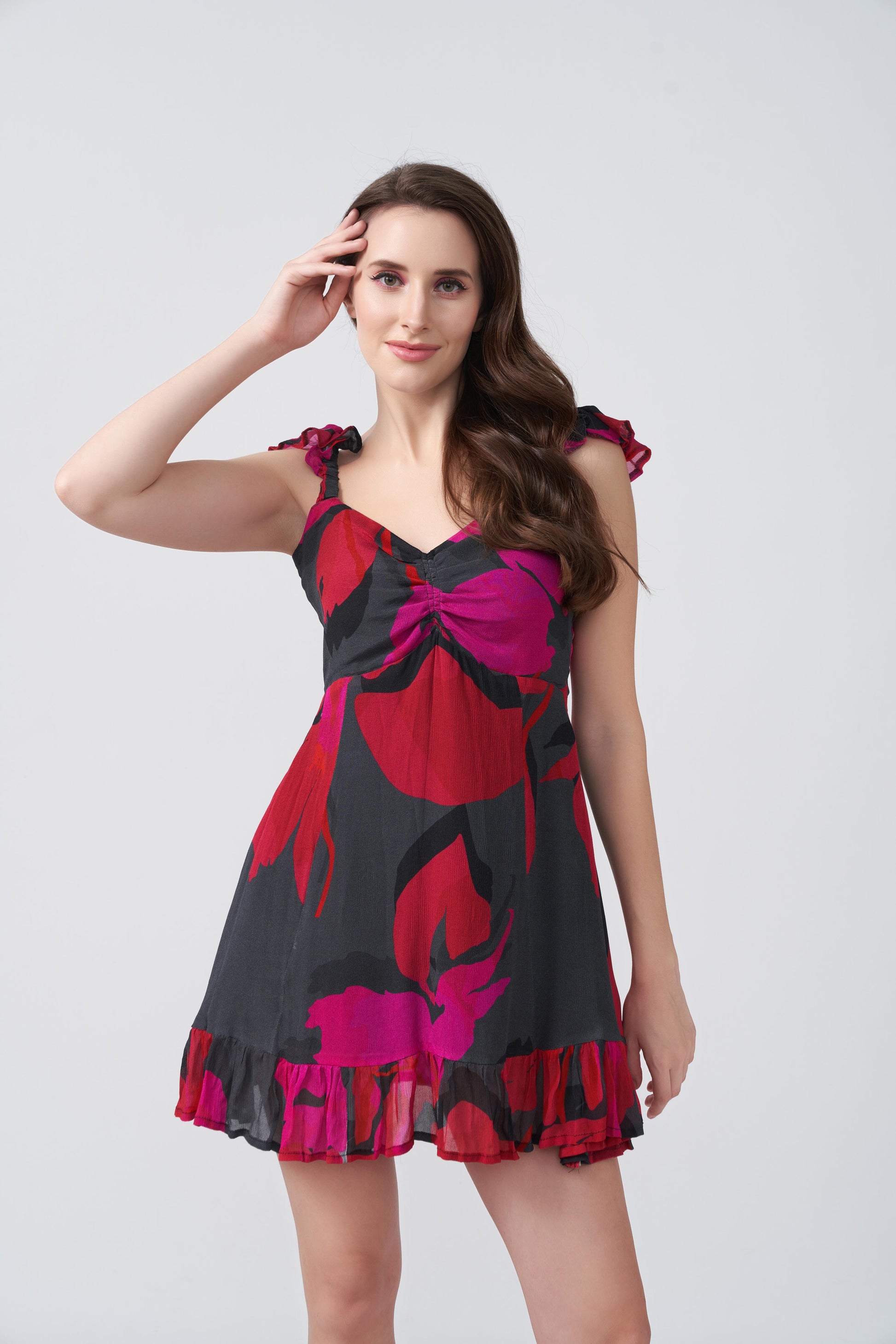 Model posing with hand in hair, wearing short red and pink floral dress with playful frill strap design.