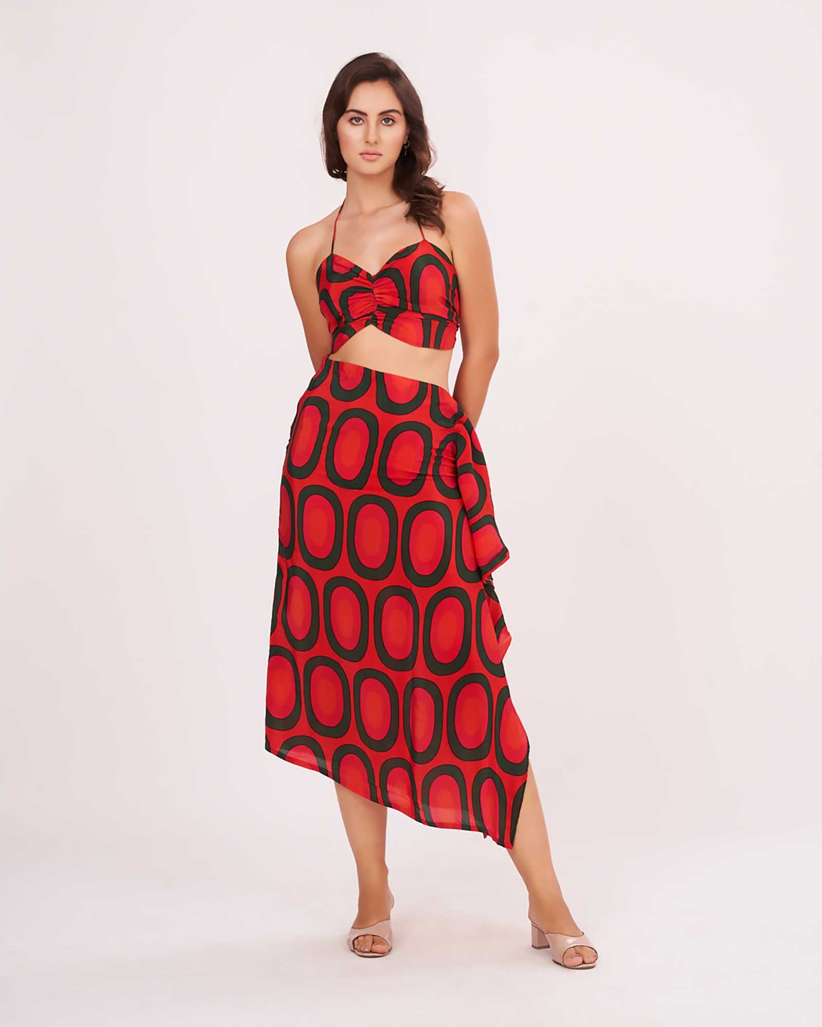 Model facing forward with both hands at her back, showcasing the red bralette top and high-waisted printed skirt with slit.