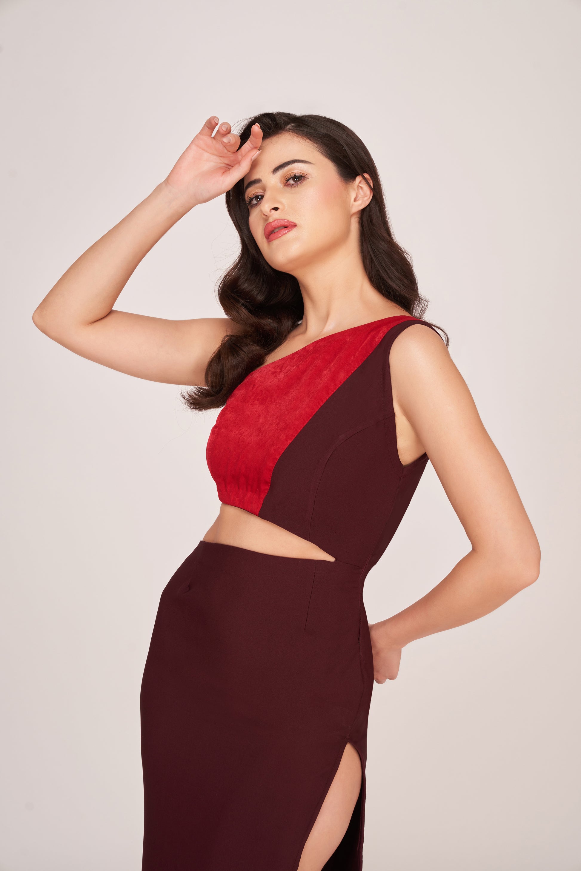 Angled view of model in brown and red panelled cut-out dress with one shoulder exposed, touching her head.