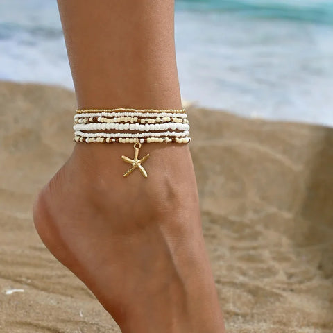 A stylish set of stacked seed bead anklets in cream, white, and gold tones featuring a dangling gold starfish charm.