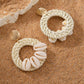 A pair of Shell Ring Drop Earrings featuring white cowrie shells and woven straw hoops resting on natural beach sand.