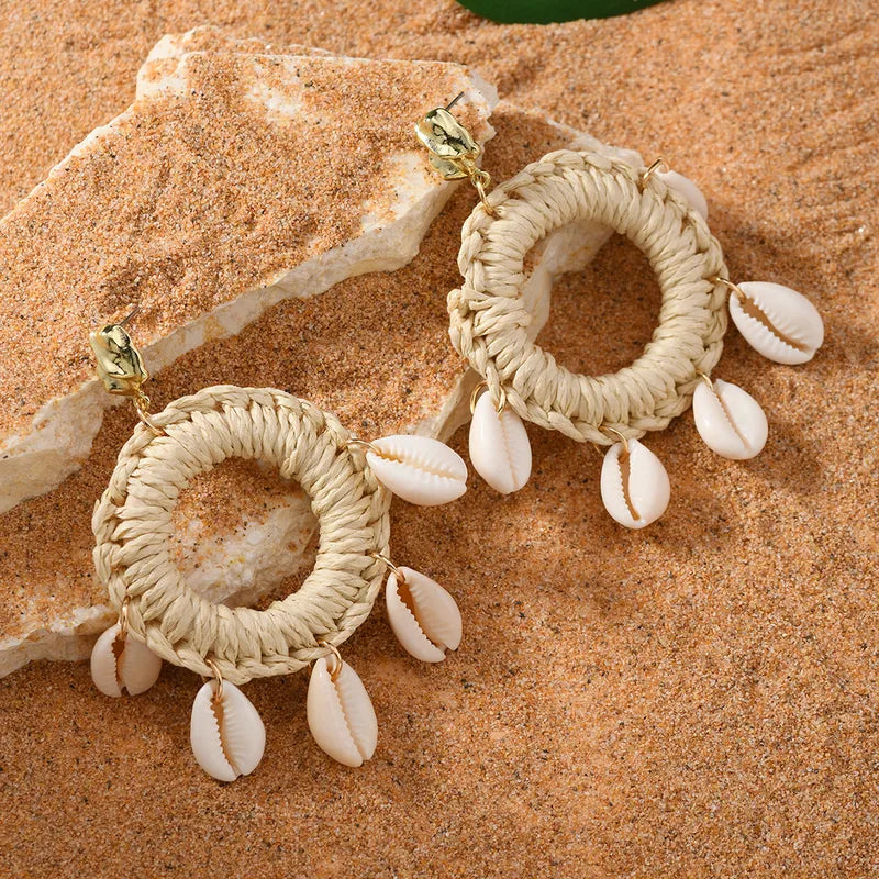 Bohemian imitation gold Shell Ring Drop Earrings featuring dangling white cowrie shells on woven straw hoops, displayed on a natural sand background.