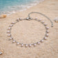 A Dainty imitation silver chain anklet with dangling white pearls laying on beach sand, perfect for summer vacations.