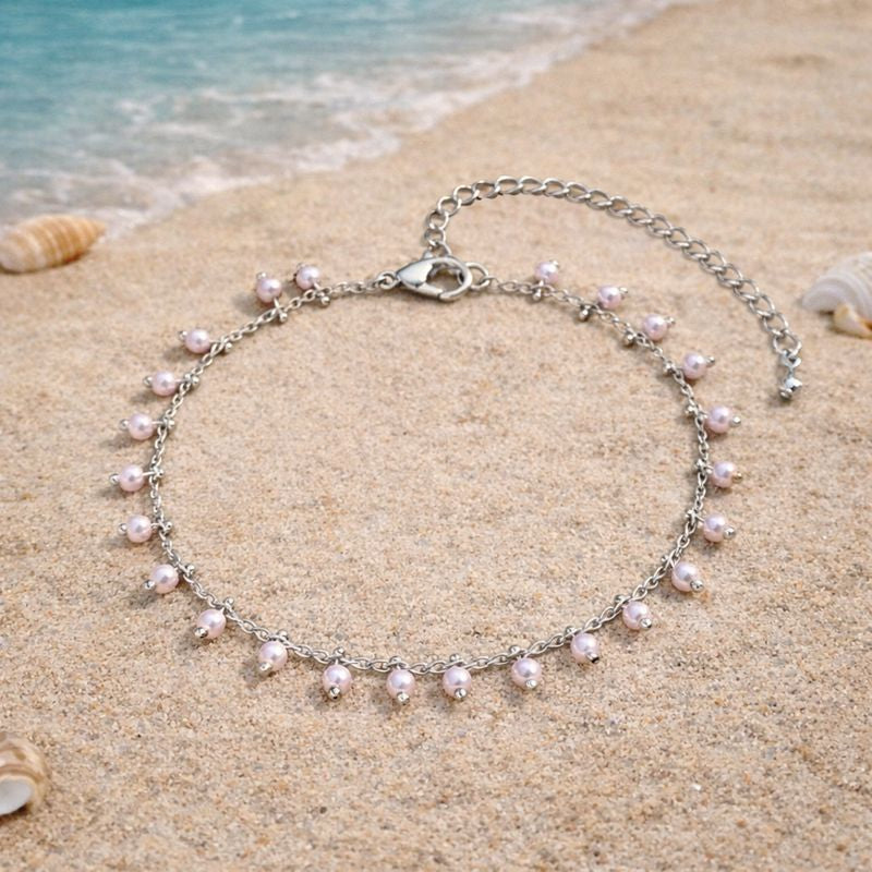 A Dainty imitation silver chain anklet with dangling white pearls laying on beach sand, perfect for summer vacations.