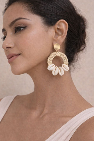A woman wearing large bohemian Shell Ring Drop Earrings featuring white cowrie shells attached to a woven straw hoop with a gold-tone stud.