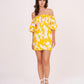 Model wearing a yellow off-shoulder mini dress with floral print and puff sleeves, standing front-facing with hands in pockets.