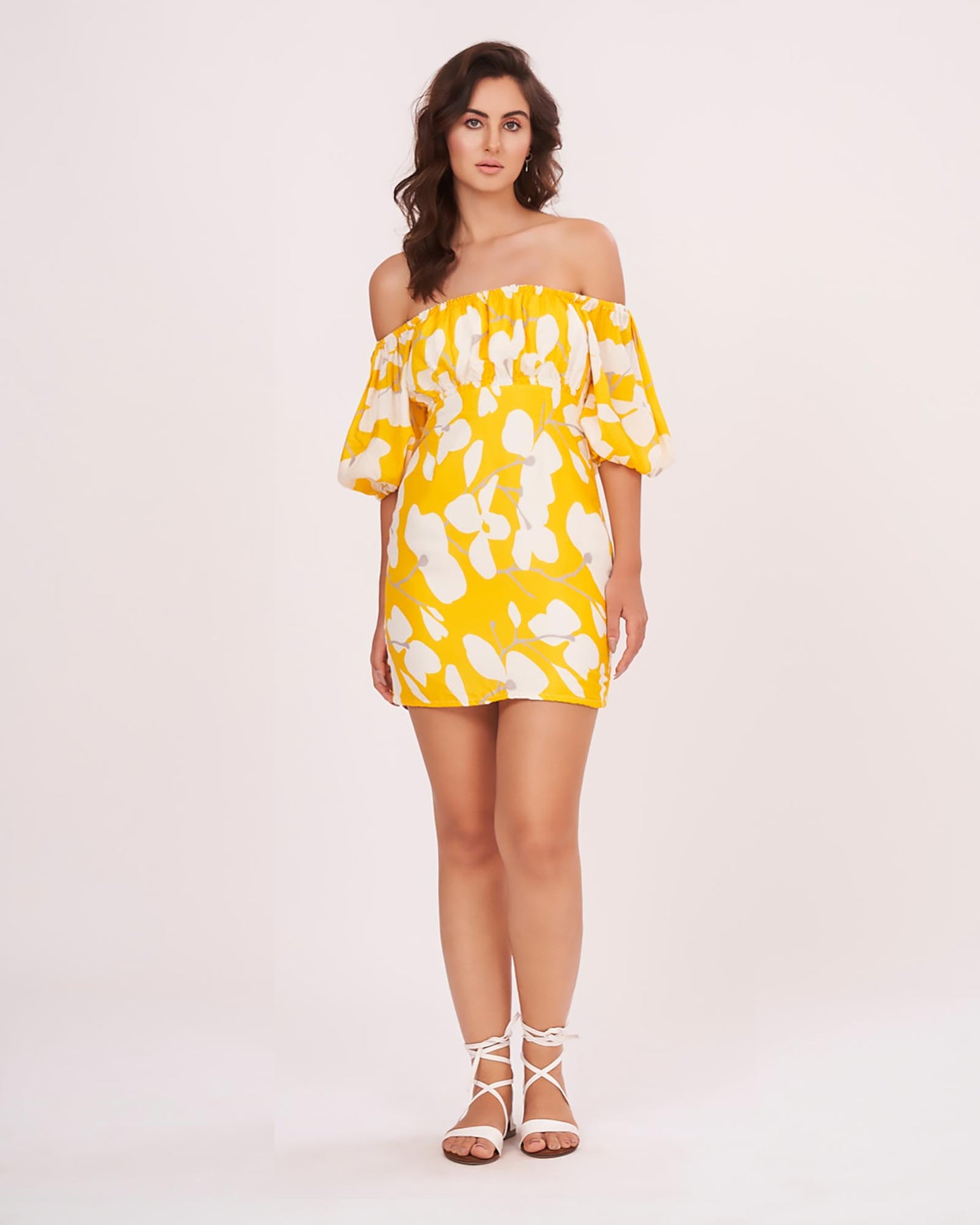 Model wearing a yellow off-shoulder mini dress with floral print and puff sleeves, standing front-facing with hands in pockets.