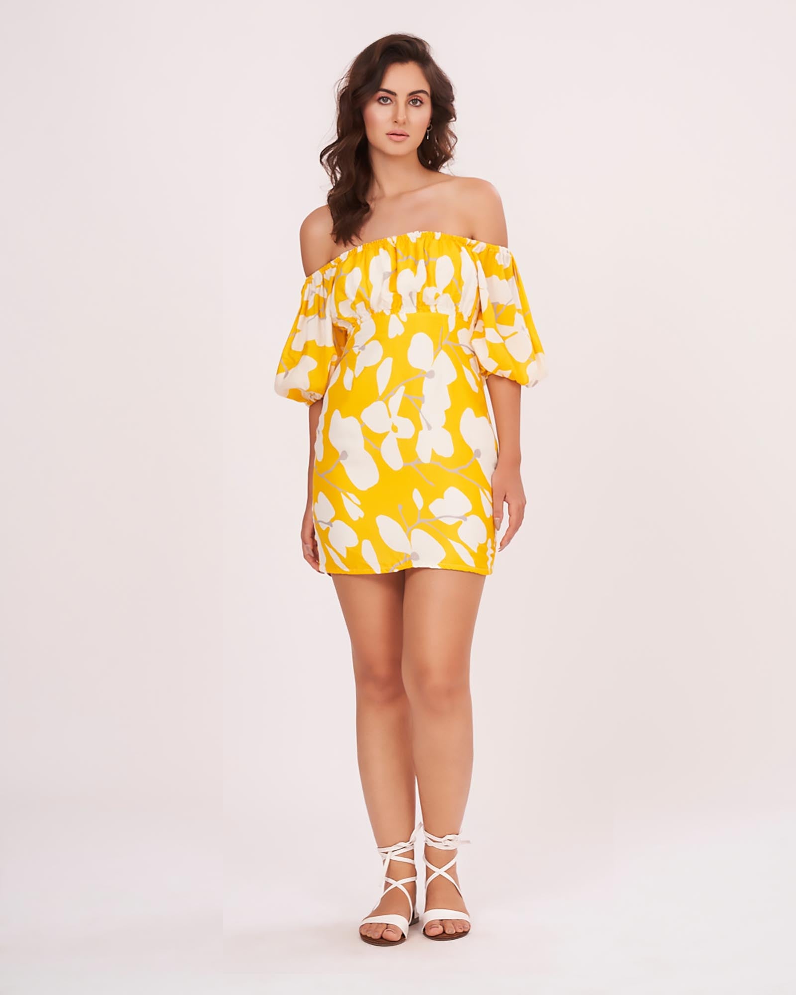 Model wearing a yellow off-shoulder mini dress with floral print and puff sleeves, standing front-facing with hands in pockets.