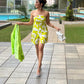 Model walking by the poolside in a vibrant yellow printed tube dress, styled with a shrug and heels.