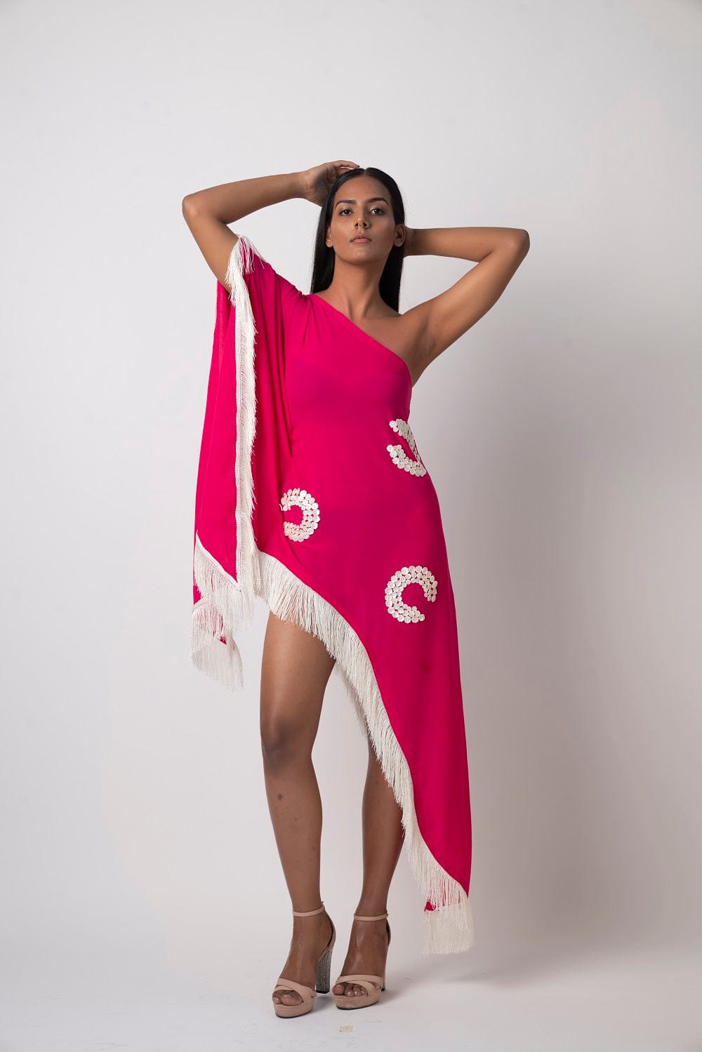 Model poses with both arms raised wearing a pink one-shoulder georgette kaftan dress adorned with shell embroidery and fringe