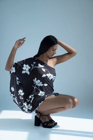 Model seated against a wall, arms raised playfully, showcasing the airy silhouette and shell-embellished hem of the black kaftan dress.