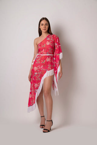 Walking pose of model in asymmetrical pink printed dress with one-shoulder design and flowing fringe hem.