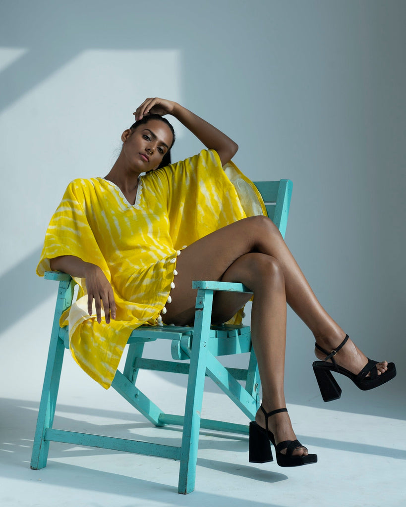 Yellow tie-dye kimono dress styled in a relaxed seated pose, model lounging on teal chair with hand on head.